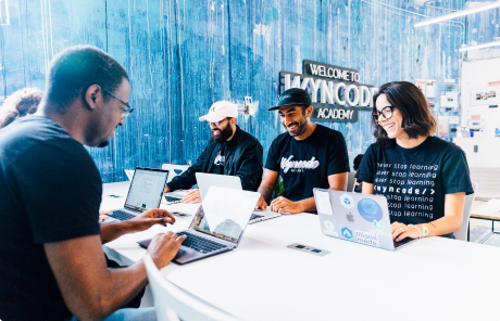 Smiling coding bootcamp students sitting together at a large table, collaborating and learning to code.